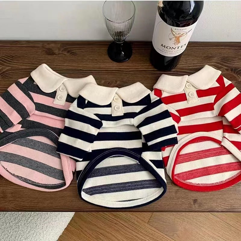 A collection of stylish striped polo shirts for dogs laid out on a wooden surface. The shirts come in three color combinations: pink with grey stripes, navy with white stripes, and red with white stripes, each featuring a polo collar with buttons and a neatly folded matching underbelly strap