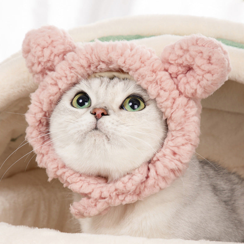 A silver tabby cat with striking green eyes peering out from beneath a fluffy pink cat hat with large, round ears, giving it a playful and adorable appearance. The cat is inside a cozy-looking, light beige pet bed, which adds to the warmth of the scene